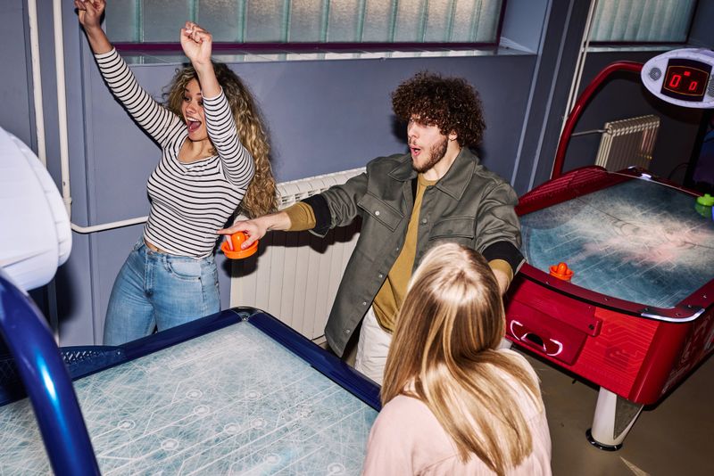 A diverse group of friends compete in an air hockey match at an indoor arcade, celebrating and laughing under dramatic direct flash lighting in a colorful, energetic game-room atmosphere.