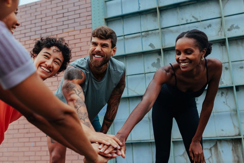 A diverse group of friends and teammates connect in a sunny outdoor setting, smiling as they place hands together in a circle. The moment captures fitness, friendship, motivation, and team spirit.