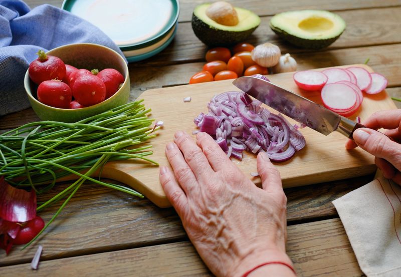 Hands chopping red onion on a wooden cutting board surrounded by avocado, tomatoes, radishes, and garlic. This rustic kitchen scene represents sustainable cooking, mindful food preparation, and the connection between plant-based ingredients and holistic wellbeing. Balanced composition with natural textures and copy space for wellness, nutrition, or sustainability messaging.
