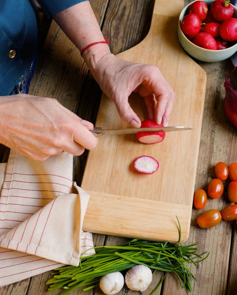 Overhead view of hands slicing a fresh radish on a wooden cutting board surrounded by herbs, tomatoes, and garlic. The scene represents mindful cooking and the use of natural ingredients that support both personal wellbeing and sustainable food practices. Rustic kitchen setting with generous copy space for healthy lifestyle or sustainability messaging.