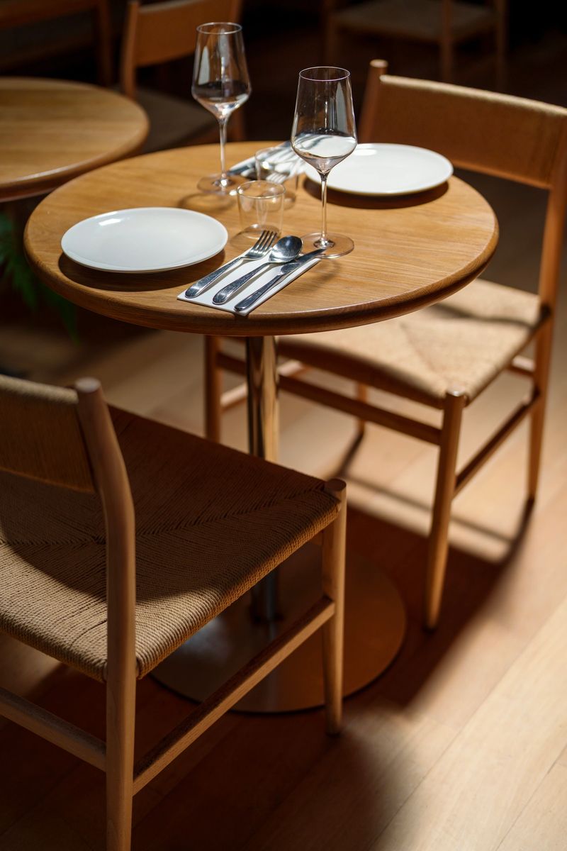 High-angle view of a minimalist round wooden table set for two in a modern restaurant. The arrangement includes white ceramic plates, polished cutlery on napkins, and elegant wine glasses, surrounded by contemporary woven chairs in warm lighting.