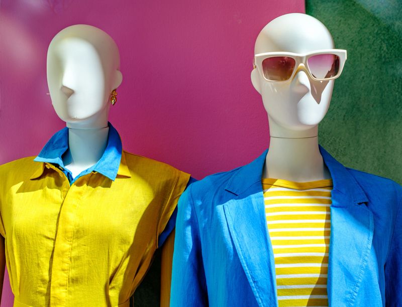 Two mannequins in a boutique wear trendy summer outfits featuring bright colors and accessories while attracting attention from passersby on a sunny day.