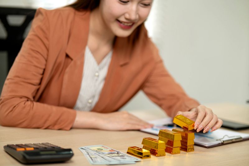 Asian adult female carefully stacks gold bars alongside currency and calculator. She plans strategic investments for future wealth financial stability.