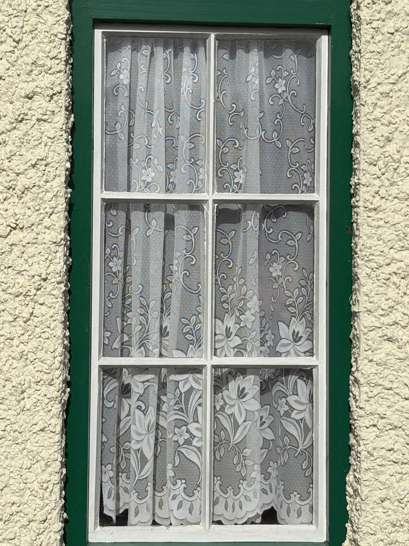 Close-up of a window with a green frame and white lace curtain against a textured exterior wall. The decorative lace fabric adds a vintage and cozy aesthetic, while the symmetrical window panes create a classic architectural detail.