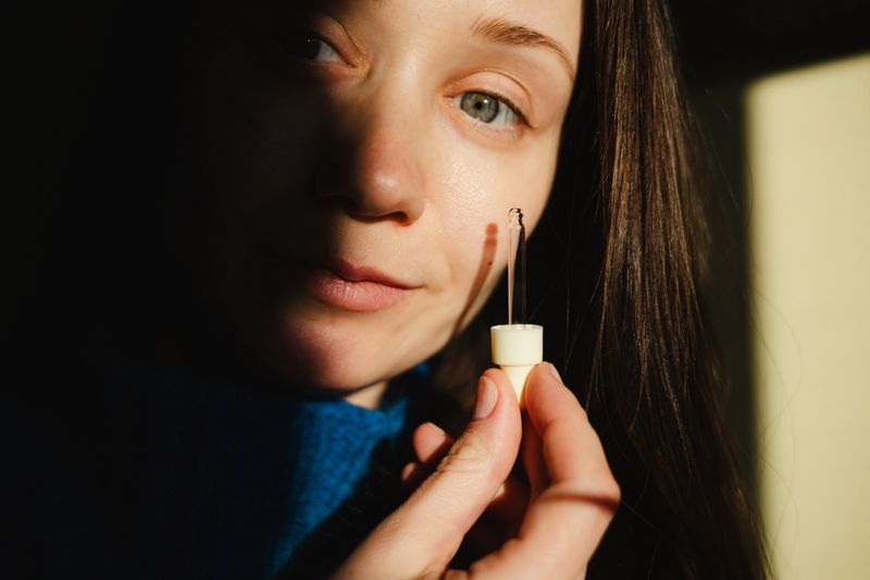 Portrait of a young woman with natural skin holding a glass pipette with a serum drop. Golden hour sunlight and shadows. Authentic beauty and skincare routine concept. Moody wellness aesthetic.