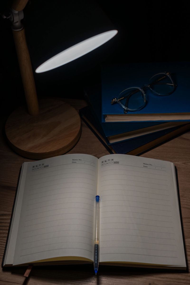 Blank notebook and pen on wooden table with books in background, illuminated by desk lamp in dark room.
