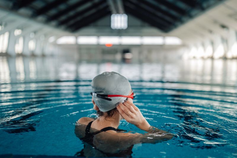Young adult swimmer adjusting goggles and swim cap at indoor pool edge, focused and ready for training or competition