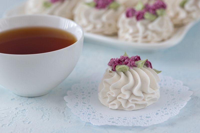 homemade Zephyr. Delicate meringue dessert topped with dried rose petals served on a lace doily beside a cup of black tea on a light blue textured surface