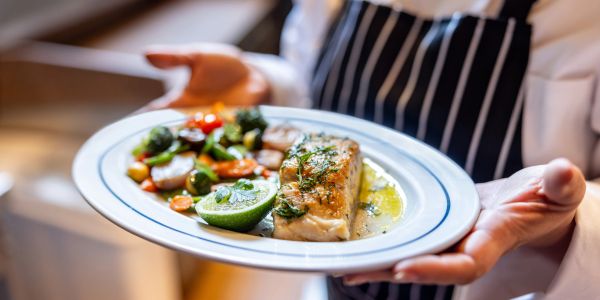 A person holds a plate of grilled fish with vegetables and lime.