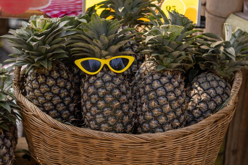 A wicker basket is filled with fresh, ripe pineapples, one of which stands out wearing bright yellow cat-eye sunglasses. This playful scene creates a fun, tropical vibe perfect for summer.