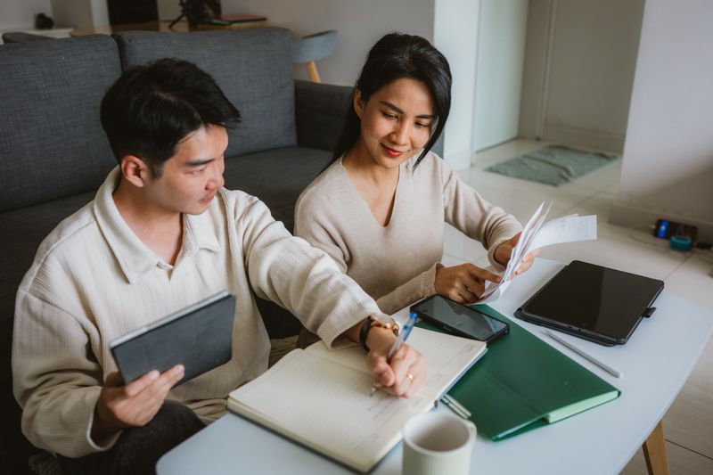 A young Asian man and woman are working together in a cozy home office, discussing taxes notes and using digital devices on a table. Preparing your Taxes concept