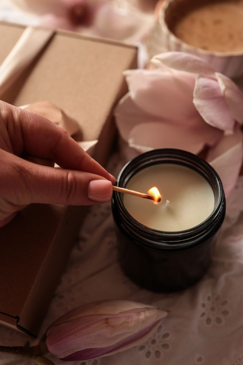 A hand ignites a candle with a match, creating a warm glow amidst delicate magnolia petals and simple gift packaging