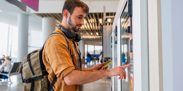 Man uses a touchscreen kiosk while holding a smartphone in a public indoor space.