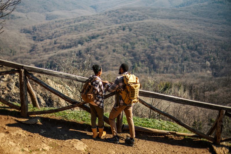 Couple with backpacks is standing on a mountain trail, enjoying the panoramic view of a valley covered with trees, embracing nature and outdoor adventure