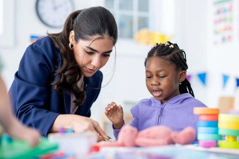 Female teacher attentively guides a young girl through a hands‑on learning activity in a bright, inclusive preschool classroom, conveying focus, encouragement, early childhood education and play‑based learning.