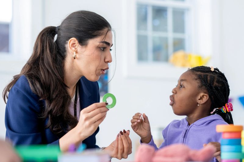 Female teacher working one-on-one with a young diverse girl in a bright classroom, using speech therapy techniques and toys to practice sounds, communication, learning and confidence building.
