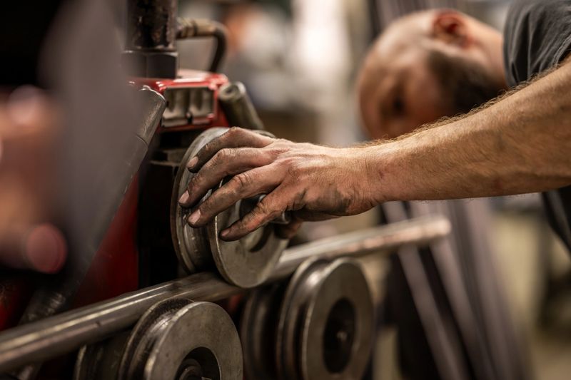 Metalworker guiding steel through roller bending machine with precise hand placement. Industrial workshop setting with metal processing equipment. Focused craftsmanship in steel fabrication work