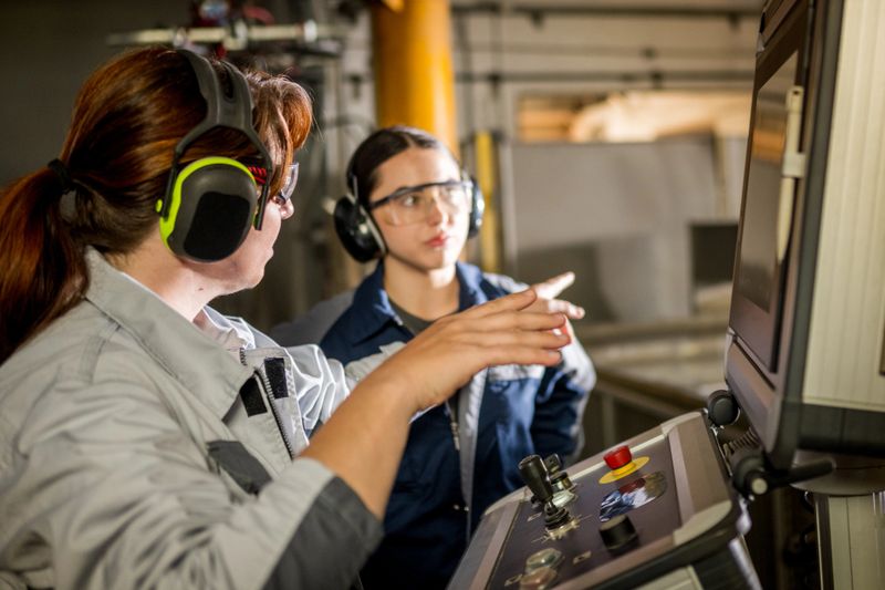 Mature and young Caucasian females wearing safety glasses and headphones, operating an industrial water cutting machine control panel in a modern manufacturing facility. The females are focused work on the machine. Focus on industrial production and focused work.