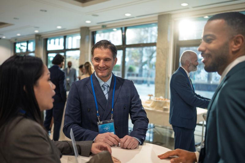 Woman and man in business attire conversing at a conference table with colleagues networking in the background. Natural light streams through large windows, creating a professional atmosphere.