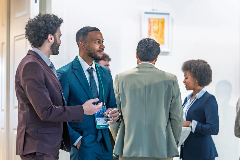 Diverse business professionals networking at a conference event, holding drinks and name tags. African American businessman and other business people socializing and talking, with bright light in the background. Business conference networking event promotes professional connections and collaborations.