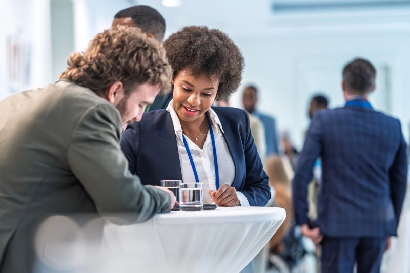 Young Hispanic businessman and multiracial businesswoman interacting at a conference event. People standing around a cocktail table, engaging in conversation and networking. The event appears to be indoors, with bright lighting and a professional atmosphere, fostering connections and exchanging ideas.