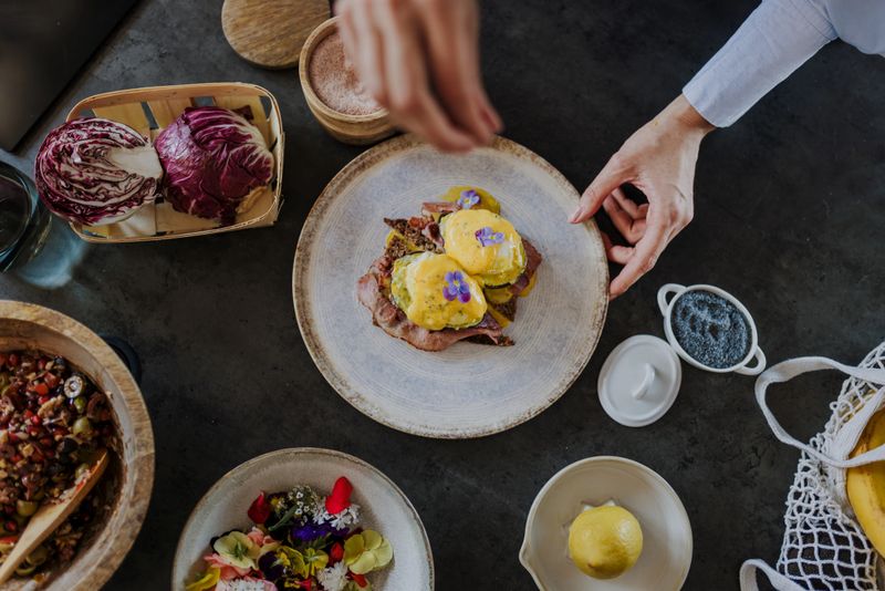 A chef meticulously garnishes a plate of Eggs Benedict, showcasing fresh ingredients like radicchio, olives, and edible flowers, ready for serving