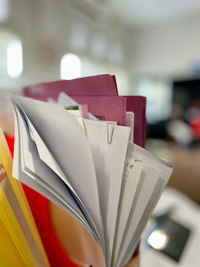 A close-up, selective focus shot of organized paper documents held together by a metal paperclip. The image shows layers of white papers and colorful folders in a bright, modern office environment. Ideal for themes related to business administration, bureaucracy, accounting, or organization.