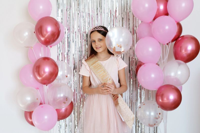 A girl wearing a dress and a sash stands among pink and gold balloons at a celebration. The backdrop has shiny decorations, creating a fun atmosphere for a special day.