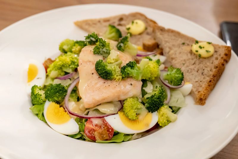 Plated salmon salad with broccoli, hard-boiled eggs, red onion, mixed greens and creamy dressing, served with buttered rustic bread on a white plate, healthy gourmet lunch or dinner closeup