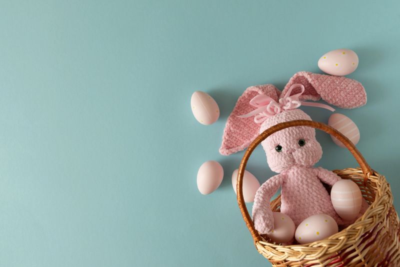 Woven basket with pink crocheted bunny doll surrounded by pastel Easter eggs against  soft blue background