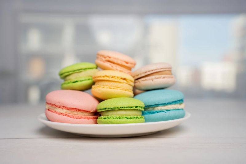 close up of colorful pastel macarons stacked on a white plate near a bright window in a minimalist kitchen or cafe setting, soft natural light, shallow depth of field and blurred background