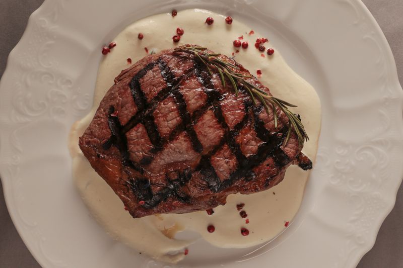 Grilled ribeye steak with creamy sauce, crosshatched char marks and rosemary sprig on white porcelain plate scattered pomegranate arils and glossy jus create festive contrast and upscale restaurant
