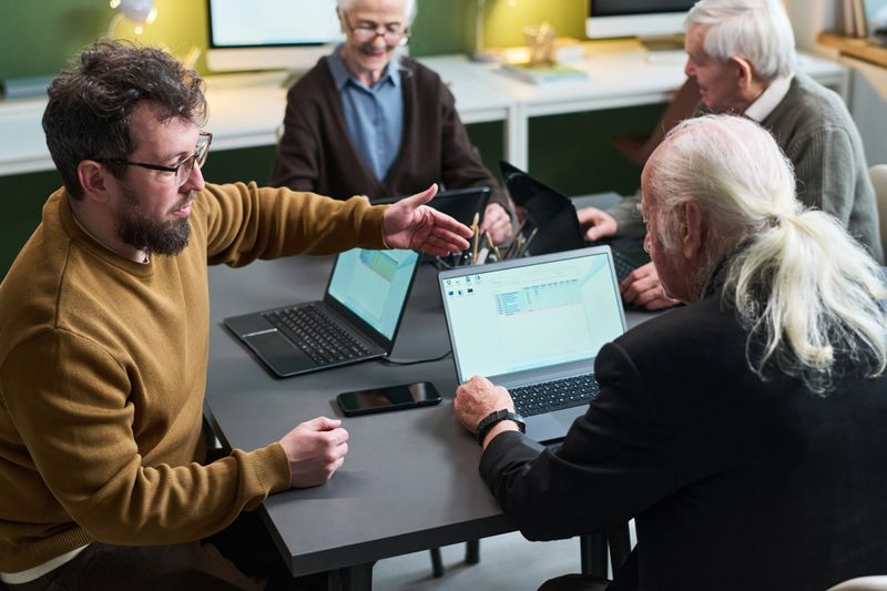 Caucasian young adult man teaching senior Caucasian man and woman computer skills during literacy course, group sitting at table using laptops, learning digital technology together