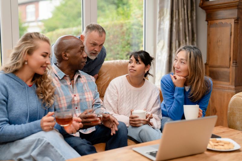 Friends sit together in a living room, enjoying warm drinks and chatting. They share laughter and stories as they relax on a couch. A laptop and cookies are on the table.