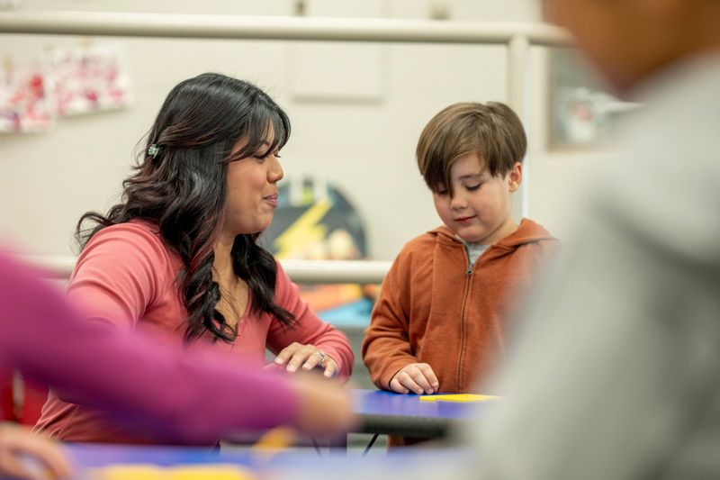 Elementary teacher guiding a young student at a classroom table, focused on hands-on learning and one-on-one support in a multicultural school environment promoting engagement and confidence.