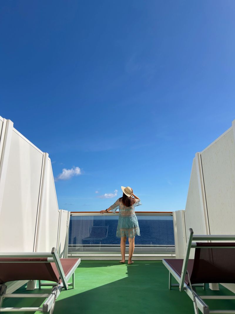 Latin american tourist female wearing a sun hat and summer dress standing on a private cruise ship balcony overlooking the vast blue ocean and sky, embracing solo travel and relaxation during a tropical vacation.She's wearing a straw hat, sunglasses, bikini and a green beach dress.