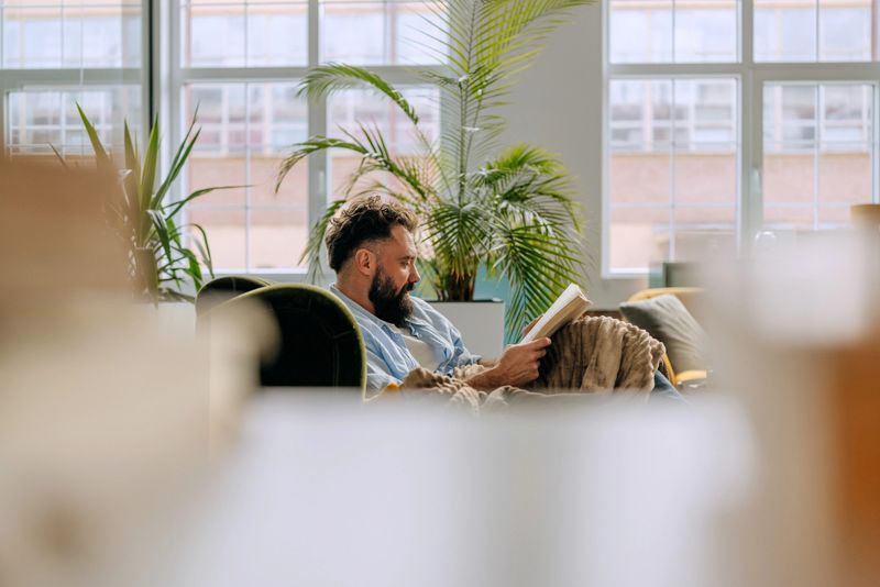 A man sits on a couch in his living room reading a book. He enjoys his weekend at home surrounded by plants and relaxed furnishings. It is a quiet moment of leisure.