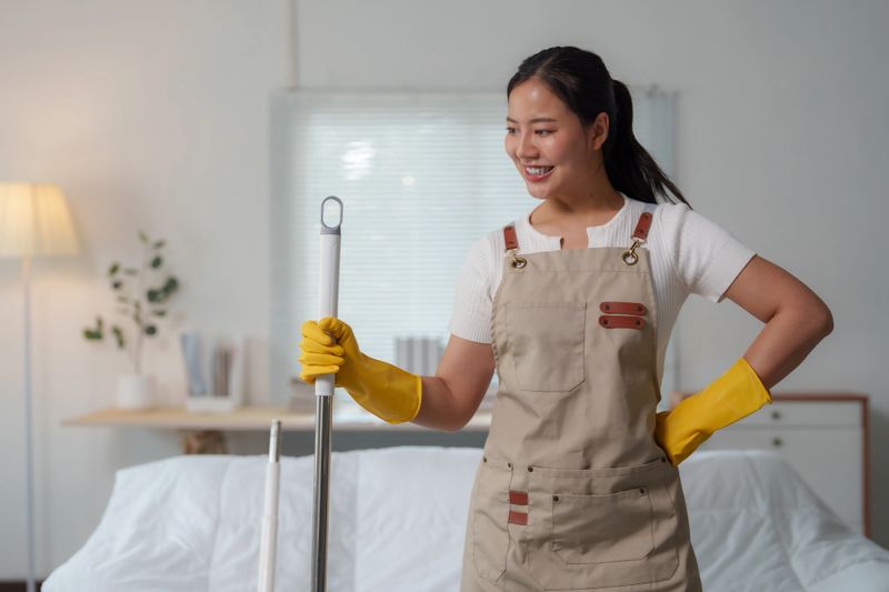 Happy housekeeper wearing yellow gloves and apron, holding cleaning mop, smiling and looking away, ready to clean bedroom in modern apartment