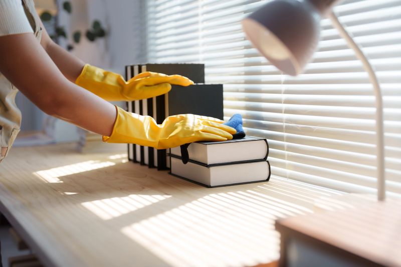 Close-up of a housekeeper wearing yellow gloves cleaning a bookshelf with a microfiber cloth, ensuring a dust-free and hygienic environment in a sunlit room