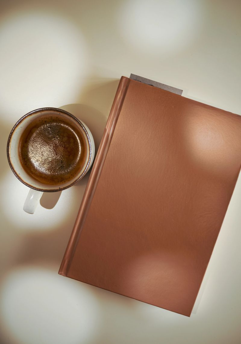 Atmospheric overhead shot of a rich terracotta hardcover book bathed in soft dappled sunlight, creating elegant shadow play across both the book cover and the cream-colored surface. A rustic speckled ceramic mug holds a dark, crema-topped espresso beside it, completing a warm, earthy composition perfect for autumn lifestyle campaigns, wellness brands, and cozy reading content.
