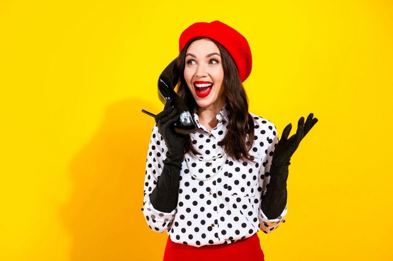 Playful retro portrait of a fashionable woman wearing a red beret and polka dot blouse, holding a high-heel shoe to her ear like a phone and smiling against a vivid yellow backdrop.