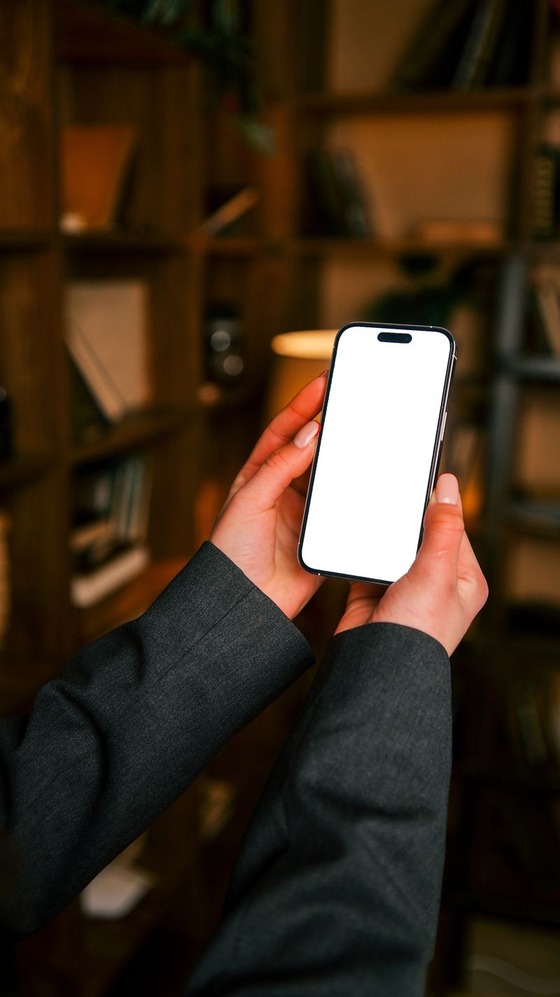 A person in a suit jacket holds a modern smartphone with a blank screen, ready for content creation or display, against a blurred background of bookshelves