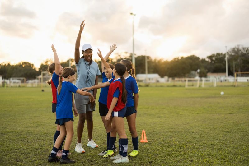 Diverse female coach and young girls huddling on a soccer field, celebrating success and showing teamwork during sports training