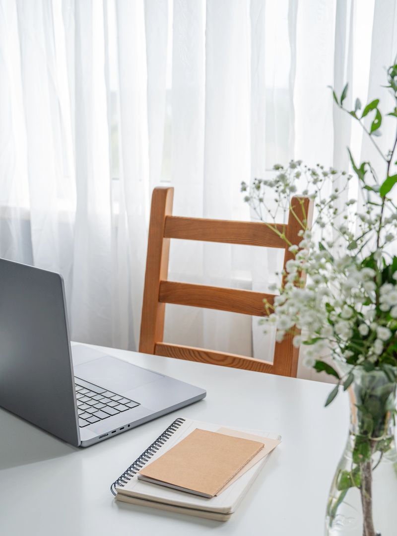 A laptop, notepad, and vase of flowers on a white table in natural light, against a wooden chair and translucent curtain. Concept for a calm and cozy workspace. Front view, copy space.