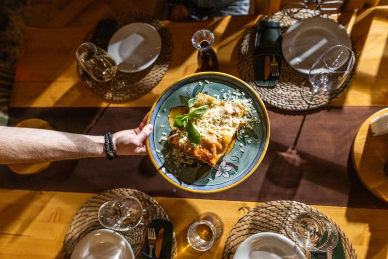 Overhead shot of a served pasta dish topped with grated cheese and basil being placed on a rustic wooden dinner table set for a family meal, warm lighting and inviting communal atmosphere.