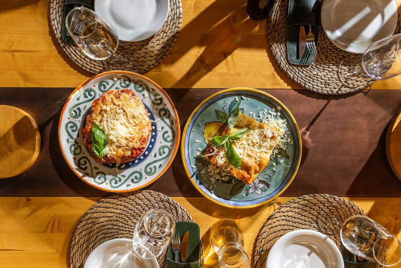 Overhead view of an Italian dinner table featuring two baked pasta dishes topped with cheese and basil, surrounded by woven placemats, glassware and warm natural light for a cozy meal scene.