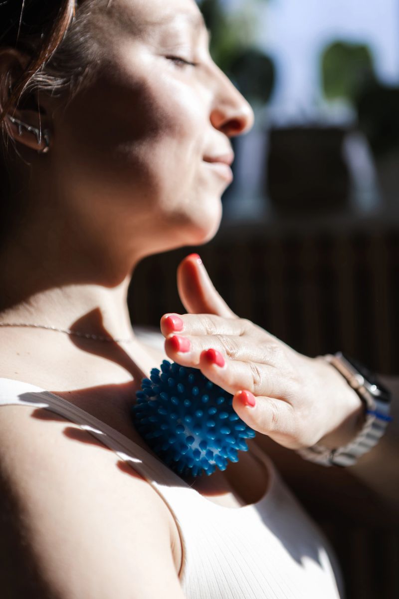 Close up of an adult woman applying a textured massage ball to the neck and shoulder area. Concept of self care, muscle relaxation, recovery, mindfulness, and home wellness routine in natural light.