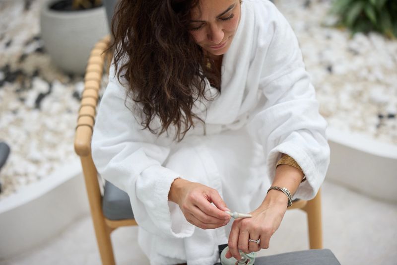 Woman wearing a white robe applies hand cream while seated, conveying self-care, relaxation and skincare routine in a calm, minimalist indoor setting with natural textures and soft lighting.
