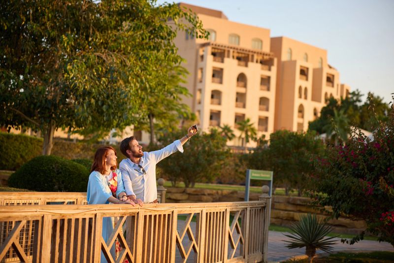 A smiling couple pause on a wooden bridge in a landscaped resort garden, taking a selfie and enjoying a relaxed, romantic vacation moment in warm late-afternoon sunlight.