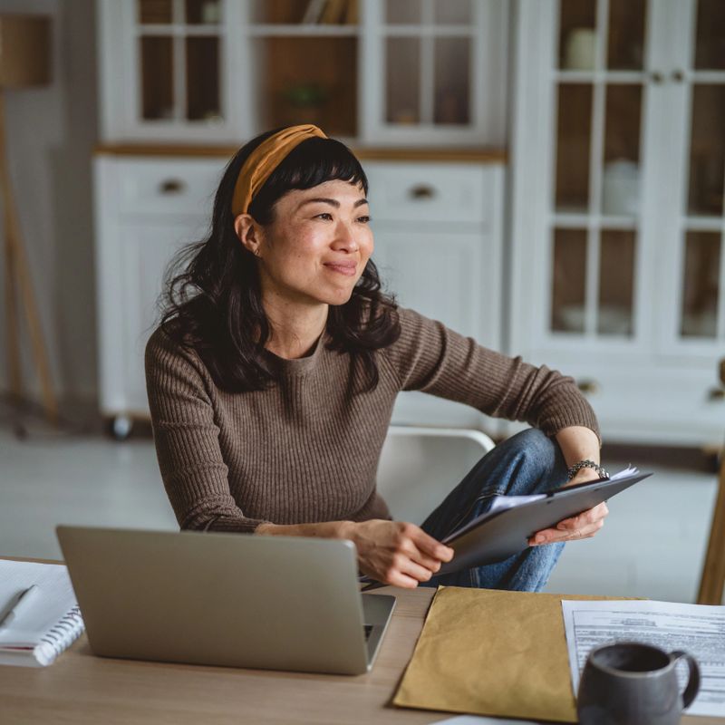 Cheerful asian woman looking away while working on a laptop from her comfortable home office, holding a clipboard with documents for her small business
